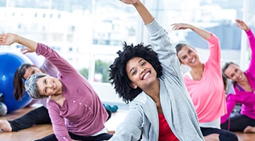 A group of women smiling at the camera as they stretch during a fitness class.