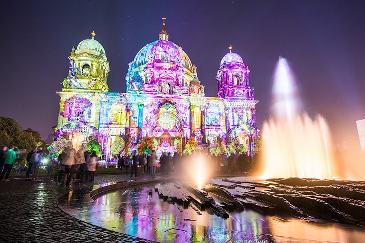 A large cathedral in Berlin which has been covered in multicolored light at night, with an outdoor fountain in the foreground.