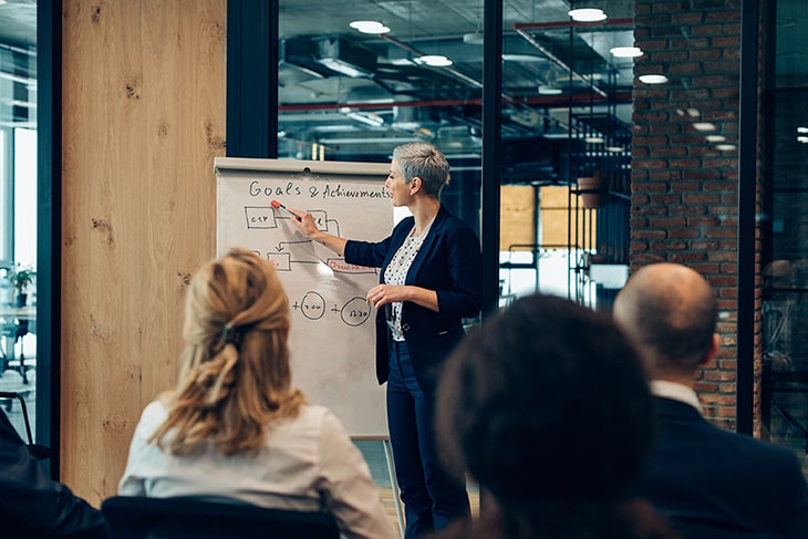 A business woman pointing to a chart drawn on a large paper as she speaks to other professionals.