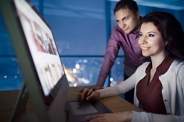 photo of a woman sitting in front a computer using a design program while a male co-worker observes.